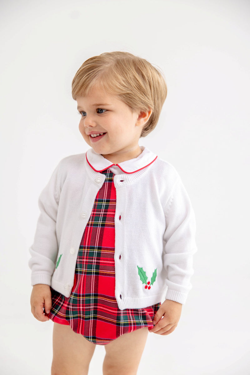 A toddler smiles and looks to the side against a plain white background, wearing The Beaufort Bonnet Company’s Cambridge Cardigan in Worth Avenue White with Holly over a red plaid dress.