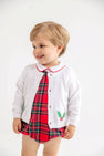 A toddler smiles and looks to the side against a plain white background, wearing The Beaufort Bonnet Company’s Cambridge Cardigan in Worth Avenue White with Holly over a red plaid dress.