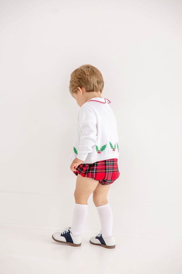 A young child stands facing a white wall, wearing The Beaufort Bonnet Company’s Cambridge Cardigan in Worth Avenue White with Holly.