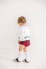 A young child stands facing a white wall, wearing The Beaufort Bonnet Company’s Cambridge Cardigan in Worth Avenue White with Holly.