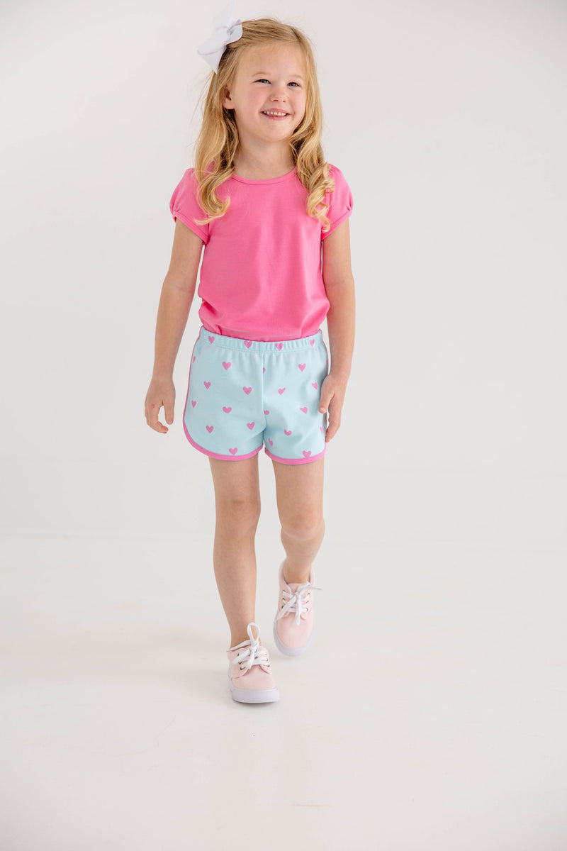 A young girl with long blonde hair and a white bow is pictured wearing The Beaufort Bonnet Company’s Penny’s Play Shirt & Onesie in Winter Park Pink, paired with light blue shorts featuring pink hearts. She walks and smiles against a plain white background, bringing a bright and cheerful touch to her everyday outfit.