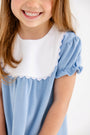 A young girl smiles while wearing The Beaufort Bonnet Company’s Pharish Frock in Barrington Blue with Worth Avenue White. The dress features puff sleeves and a white scalloped collar. Her face is partially out of frame, and her long brown hair falls loosely over her shoulders.