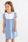 A young girl with long brown hair wears The Beaufort Bonnet Company’s Pharish Frock in Barrington Blue with Worth Avenue White, featuring puff sleeves and a large white scalloped collar. She smiles and looks to the side against a plain white background.