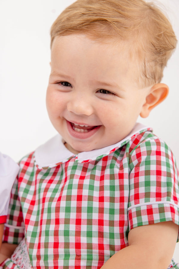A smiling young child with light brown hair is wearing The Beaufort Bonnet Company’s Brentley Bubble in Richmond Red & Kiawah Kelly Green Gingham—a festive holiday baby outfit with a white collar—while sitting against a plain white background.