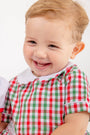 A smiling young child with light brown hair is wearing The Beaufort Bonnet Company’s Brentley Bubble in Richmond Red & Kiawah Kelly Green Gingham—a festive holiday baby outfit with a white collar—while sitting against a plain white background.