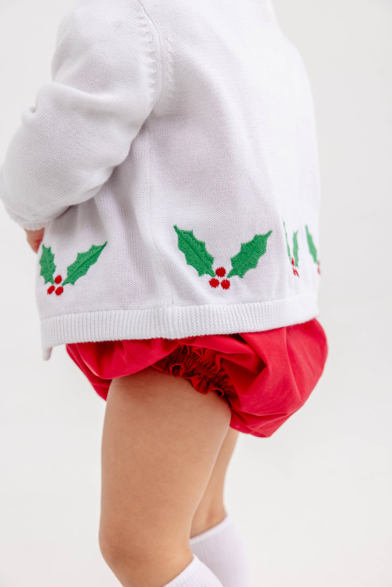 A toddler wearing The Beaufort Bonnet Company’s Cambridge Cardigan in Worth Avenue White with Holly—featuring green holly leaf designs and red berries—paired with red bloomers and white socks, stands against a light background.