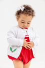 A toddler with curly hair and a white bow wears The Beaufort Bonnet Company’s Cambridge Cardigan in Worth Avenue White with Holly over a red dress, looking down with hands on their stomach against a white background.
