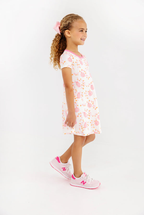 A young girl with curly hair, wearing a pink hair bow and The Beaufort Bonnet Company’s Polly Play Dress - Gourd-geous (Palmetto Pearl) with Romany Rose crafted from soft Pima Cotton, paired with pink and white sneakers, stands smiling against a plain white background.