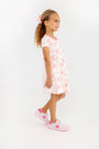 A young girl with curly hair, wearing a pink hair bow and The Beaufort Bonnet Company’s Polly Play Dress - Gourd-geous (Palmetto Pearl) with Romany Rose crafted from soft Pima Cotton, paired with pink and white sneakers, stands smiling against a plain white background.