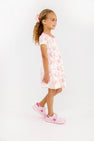A young girl with curly hair, wearing a pink hair bow and The Beaufort Bonnet Company’s Polly Play Dress - Gourd-geous (Palmetto Pearl) with Romany Rose crafted from soft Pima Cotton, paired with pink and white sneakers, stands smiling against a plain white background.