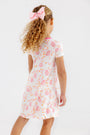 A young girl with curly hair, wearing a large pink bow and The Beaufort Bonnet Company’s Polly Play Dress – Gourd-geous (Palmetto Pearl) with Romany Rose, stands facing away against a plain white background.