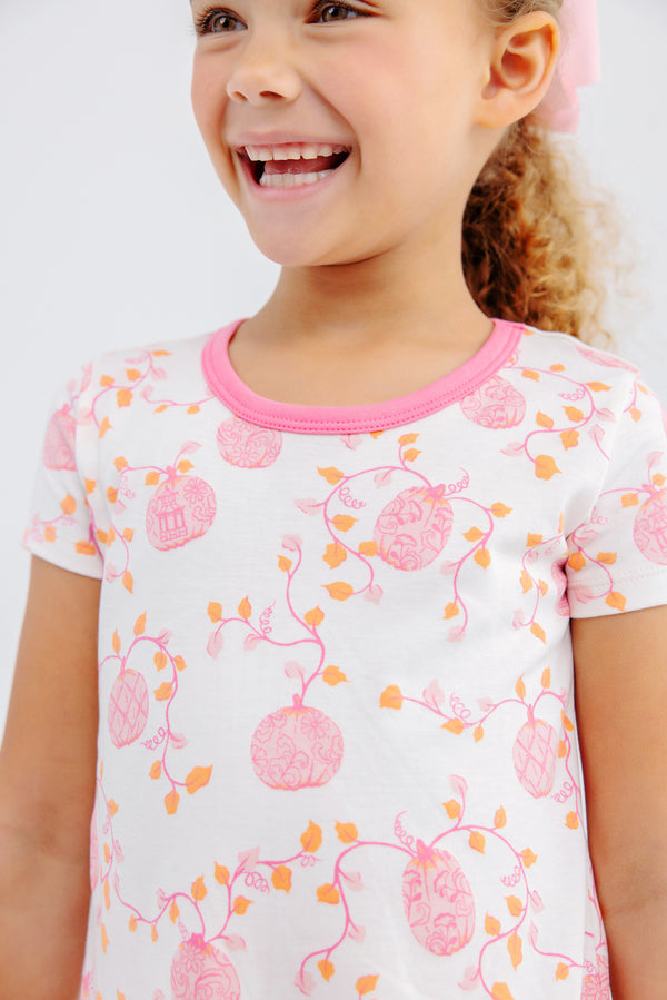 A young child wearing The Beaufort Bonnet Company’s Polly Play Dress - Gourd-geous (Palmetto Pearl) with Romany Rose beams brightly, showing their teeth, against a plain, light-colored background.