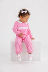 A toddler wearing a Hamptons Hot Pink Rigsby Romper with white bow smocking detail and a white collar, sitting on a white cube.