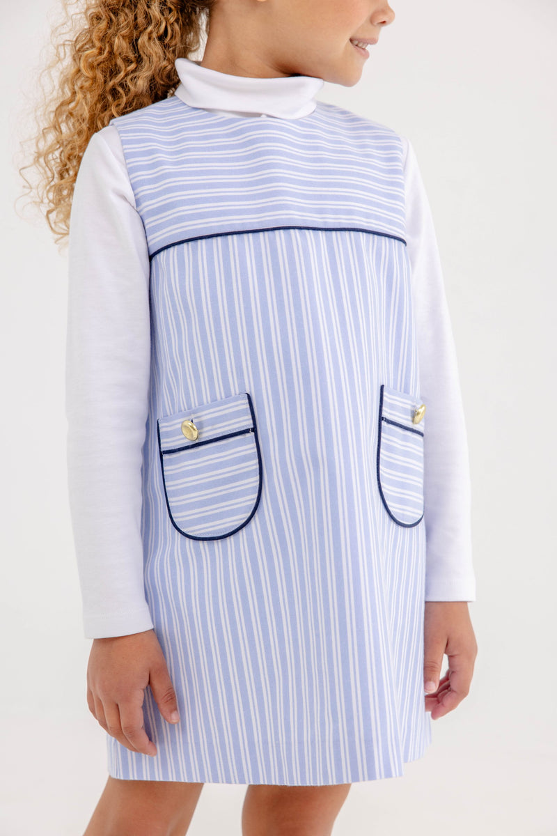 A child wears a white long-sleeve shirt under a Jill Jumper - Park City Periwinkle Stripe with Nantucket Navy, a light blue and white striped dress featuring two front pockets with navy trim and gold buttons.