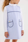 A child wears a white long-sleeve shirt under a Jill Jumper - Park City Periwinkle Stripe with Nantucket Navy, a light blue and white striped dress featuring two front pockets with navy trim and gold buttons.
