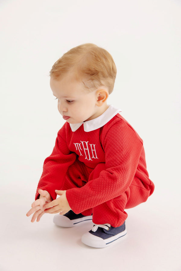 A toddler squats wearing a Potter’s Playsuit - Quilted Richmond Red with Worth Avenue White, a red quilted long-sleeved outfit with a white collar and monogramming, paired with navy blue sneakers.