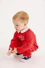 A toddler squats wearing a Potter’s Playsuit - Quilted Richmond Red with Worth Avenue White, a red quilted long-sleeved outfit with a white collar and monogramming, paired with navy blue sneakers.
