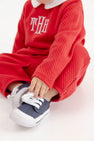 A child wears a red quilted Potter’s Playsuit - Quilted Richmond Red with Worth Avenue White, featuring a white collar and an embroidered monogram, paired with navy blue canvas sneakers as a festive Christmas baby outfit.