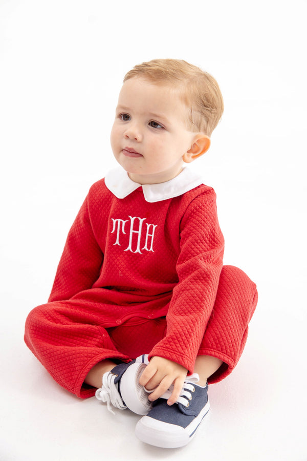 A toddler wearing a Potter’s Playsuit - Quilted Richmond Red with Worth Avenue White, featuring a red quilted design, white collar, and white monogram, sitting and holding their blue and white sneakers.