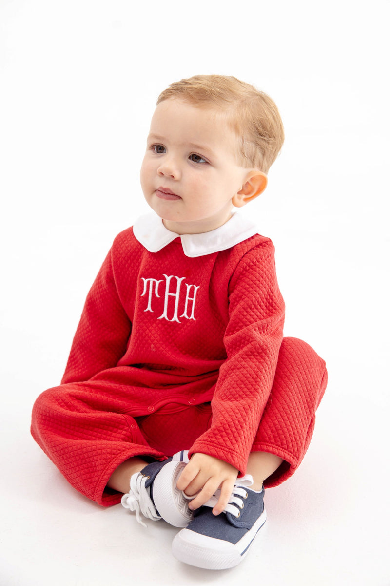A toddler wearing a Potter’s Playsuit - Quilted Richmond Red with Worth Avenue White, featuring a red quilted design, white collar, and white monogram, sitting and holding their blue and white sneakers.