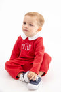 A toddler wearing a Potter’s Playsuit - Quilted Richmond Red with Worth Avenue White, featuring a red quilted design, white collar, and white monogram, sitting and holding their blue and white sneakers.
