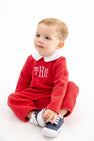 A toddler wearing a Potter’s Playsuit - Quilted Richmond Red with Worth Avenue White, featuring a red quilted design, white collar, and white monogram, sitting and holding their blue and white sneakers.