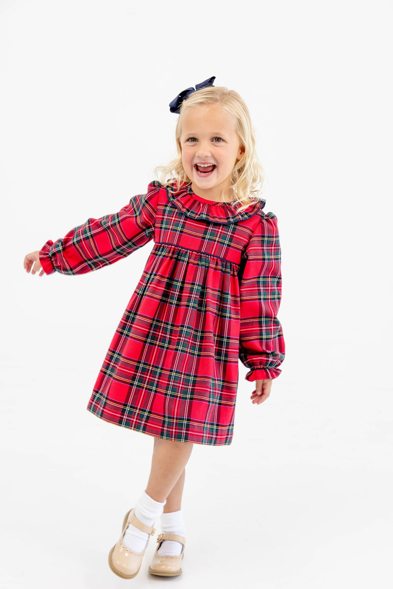 A young girl wears a Long Sleeve Ramona Ruffle Dress - Society Prep Plaid with Worth Avenue White, featuring a ruffled collar and white trim, paired with white socks and neutral Mary Jane shoes.