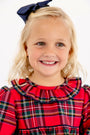 A young girl wearing a navy blue hair bow and a Long Sleeve Ramona Ruffle Dress - Society Prep Plaid with Worth Avenue White featuring a red plaid pattern and a ruffled collar.