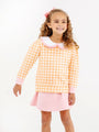 A smiling young girl with curly hair wears a pink bow, an orange and white gingham top with a white scalloped collar, and The Beaufort Bonnet Company’s Searcy Skort in Sandpearl Pink against a plain white background.