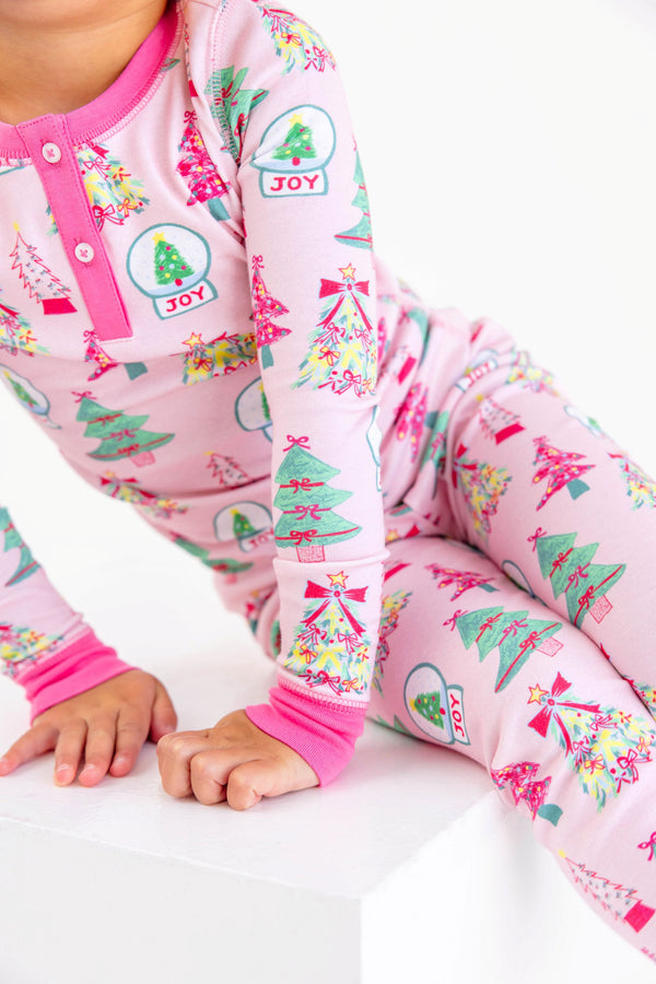 A child wearing a Payton Pajama Set - Christmas Coloring Contest with Winter Park Pink, featuring pink pajamas patterned with colorful trees and snow globes labeled JOY.
