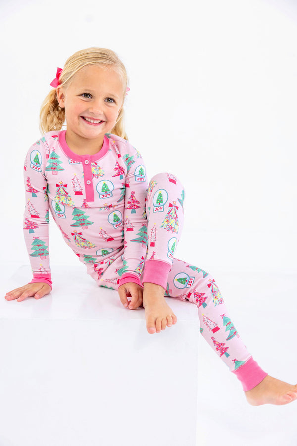 A young girl smiles while sitting on a white cube, wearing a Payton Pajama Set - Christmas Coloring Contest with Winter Park Pink, a light pink cotton pajama set patterned with Christmas trees and JOY ornaments.