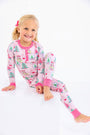 A young girl smiles while sitting on a white cube, wearing a Payton Pajama Set - Christmas Coloring Contest with Winter Park Pink, a light pink cotton pajama set patterned with Christmas trees and JOY ornaments.