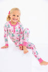 A young girl smiles while sitting on a white cube, wearing a Payton Pajama Set - Christmas Coloring Contest with Winter Park Pink, a light pink cotton pajama set patterned with Christmas trees and JOY ornaments.
