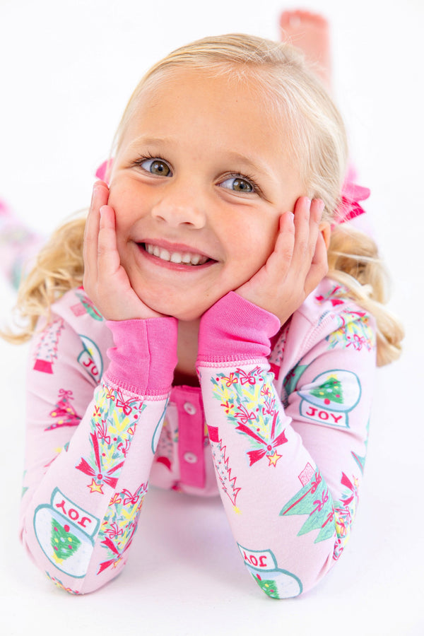 A young girl smiles with her hands on her cheeks, wearing a light pink Payton Pajama Set - Christmas Coloring Contest with Winter Park Pink, decorated with Christmas tree and snow globe illustrations.