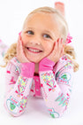 A young girl smiles with her hands on her cheeks, wearing a light pink Payton Pajama Set - Christmas Coloring Contest with Winter Park Pink, decorated with Christmas tree and snow globe illustrations.