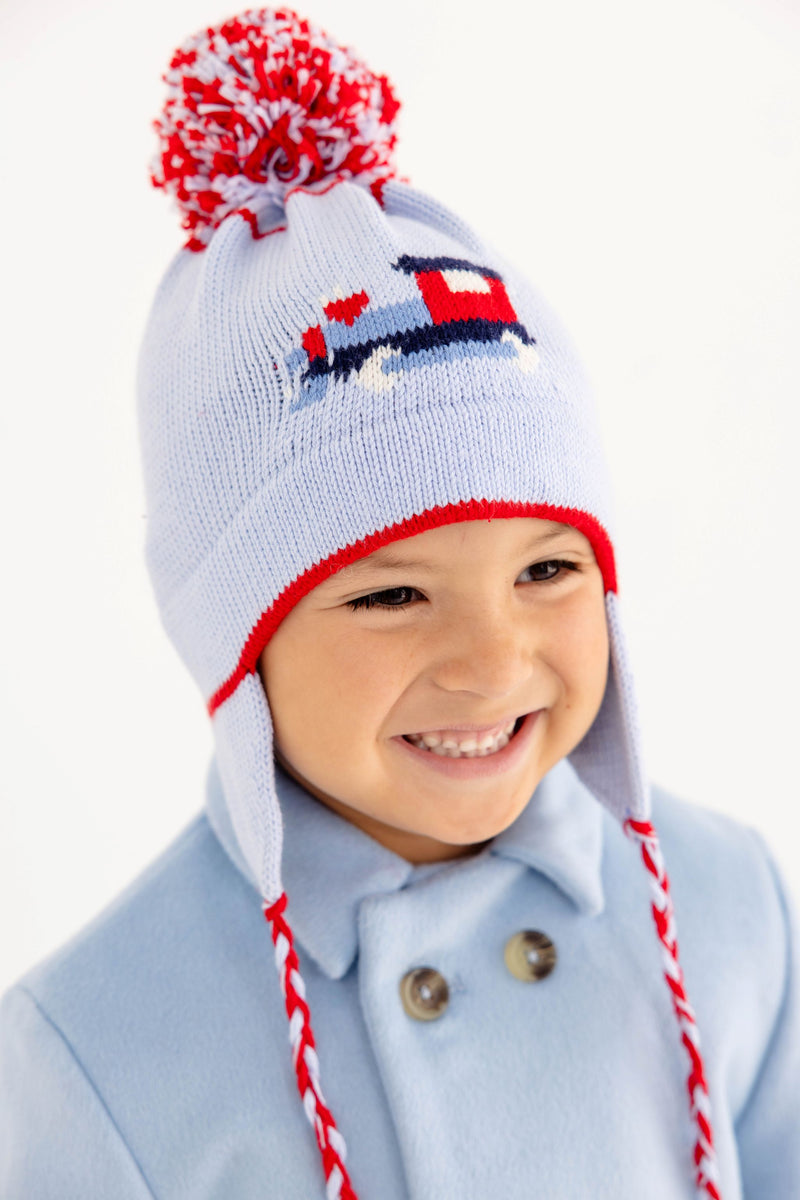 A smiling child wears The Beaufort Bonnet Company’s Parrish Pom Pom Hat in Beale Street Blue with a playful train motif, complete with ear flaps, red trim, and a red-and-white pompom.