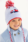 A smiling child wears The Beaufort Bonnet Company’s Parrish Pom Pom Hat in Beale Street Blue with a playful train motif, complete with ear flaps, red trim, and a red-and-white pompom.