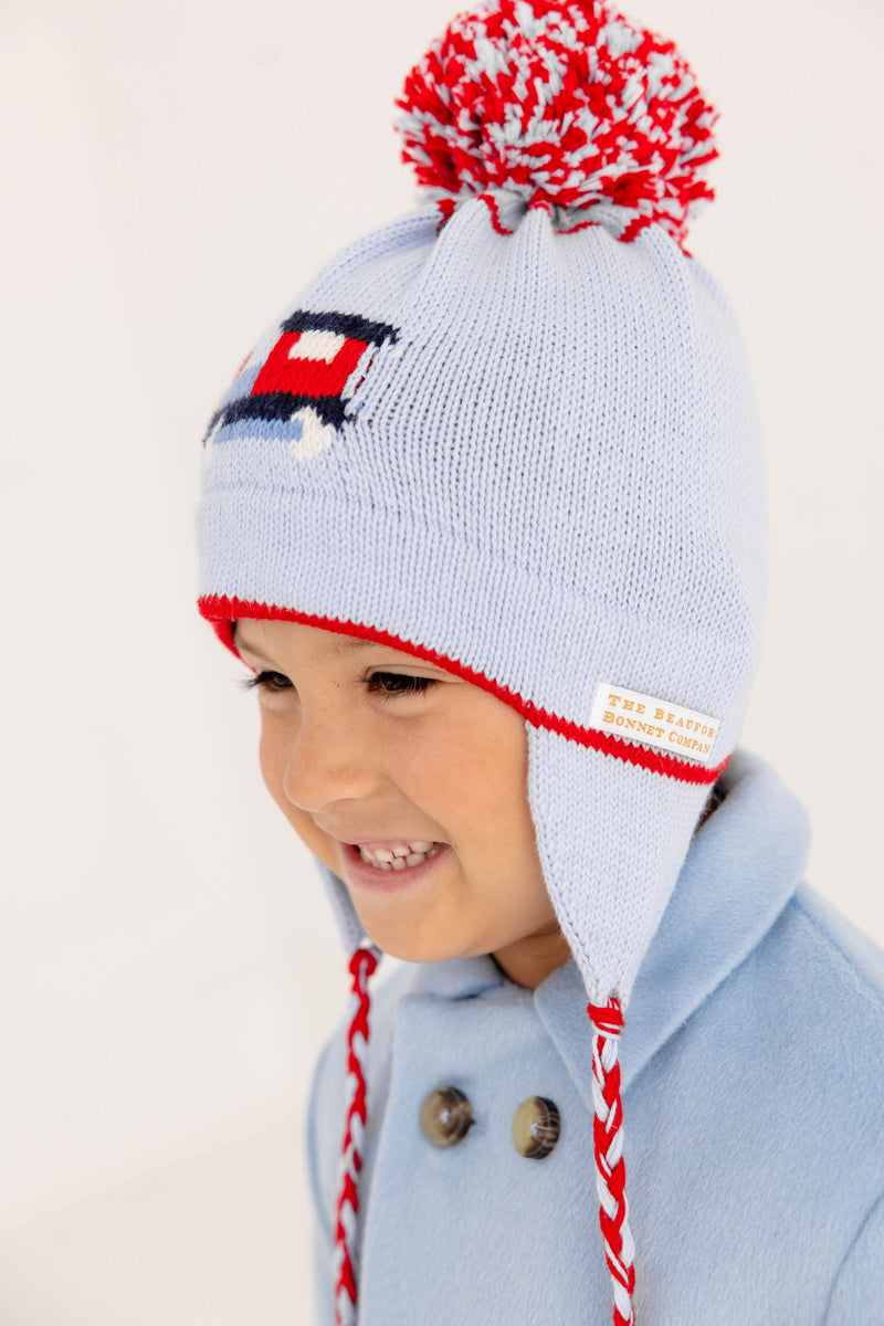 A smiling child in a light blue coat wears The Beaufort Bonnet Company’s Parrish Pom Pom Hat in Beale Street Blue with a playful train motif, featuring red, white, and blue cable-knit details, ear flaps, braided tassels, and a cheerful red pom-pom on top.