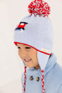 A smiling child in a light blue coat wears The Beaufort Bonnet Company’s Parrish Pom Pom Hat in Beale Street Blue with a playful train motif, featuring red, white, and blue cable-knit details, ear flaps, braided tassels, and a cheerful red pom-pom on top.