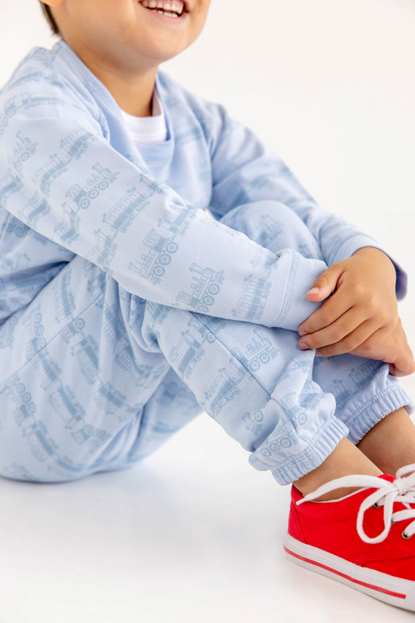 A smiling child sits with arms around their knees, wearing The Beaufort Bonnet Company’s Cassidy Comfy Crewneck in Blue Choo Choos—paired with bright red sneakers for the perfect cozy travel outfit against a white background.