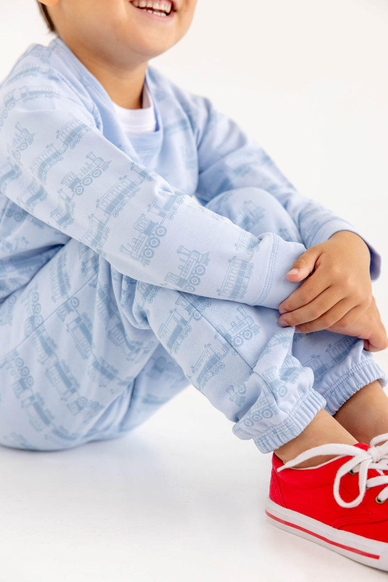 A smiling child sits with arms around their knees, wearing The Beaufort Bonnet Company’s Cassidy Comfy Crewneck in Blue Choo Choos—paired with bright red sneakers for the perfect cozy travel outfit against a white background.