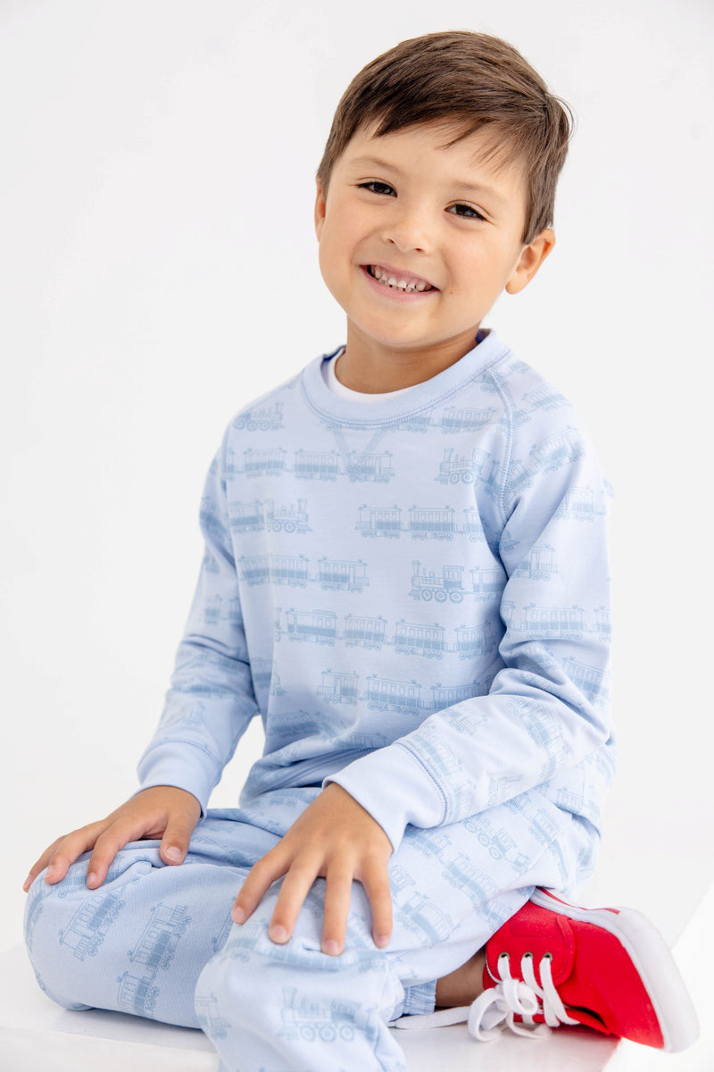 A smiling young boy with short brown hair sits against a white background, wearing The Beaufort Bonnet Company’s Cassidy Comfy Crewneck in the Blue Choo Choos print and light blue pajamas, paired with bright red sneakers.