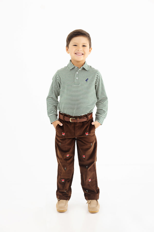 A smiling young boy stands with his hands in the pockets of his brown pants, wearing a green and white striped long-sleeve shirt, beige shoes, and The Beaufort Bonnet Company’s Bullock Belt (Leather) in Tan, against a plain white background.