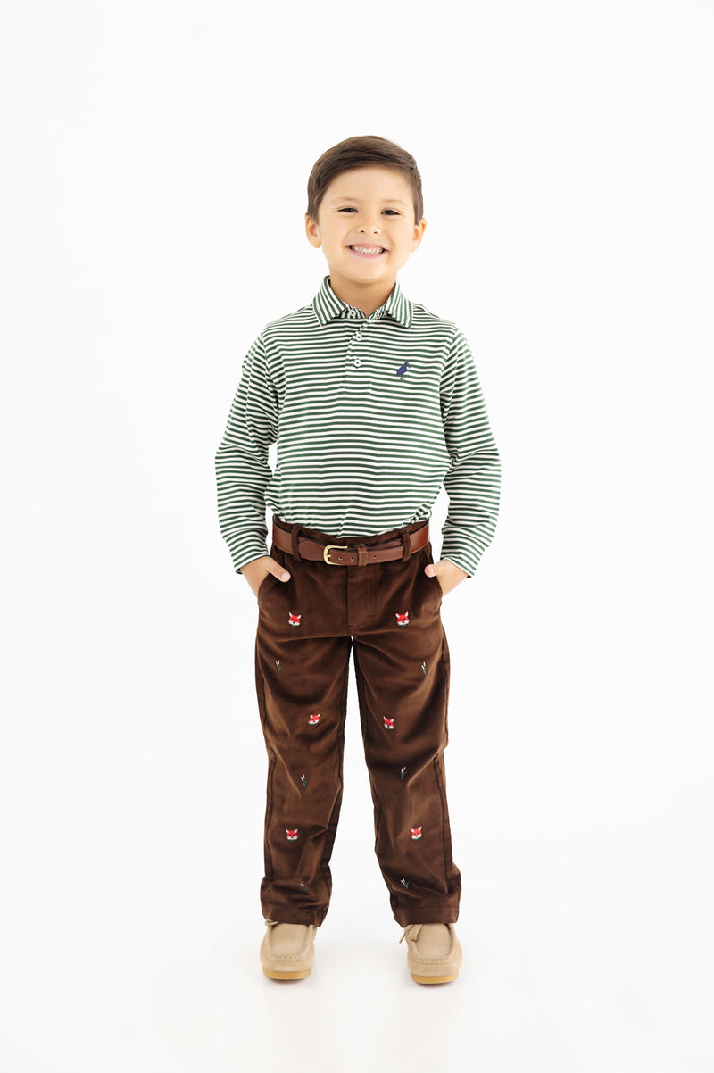 A smiling young boy stands with his hands in the pockets of his brown pants, wearing a green and white striped long-sleeve shirt, beige shoes, and The Beaufort Bonnet Company’s Bullock Belt (Leather) in Tan, against a plain white background.