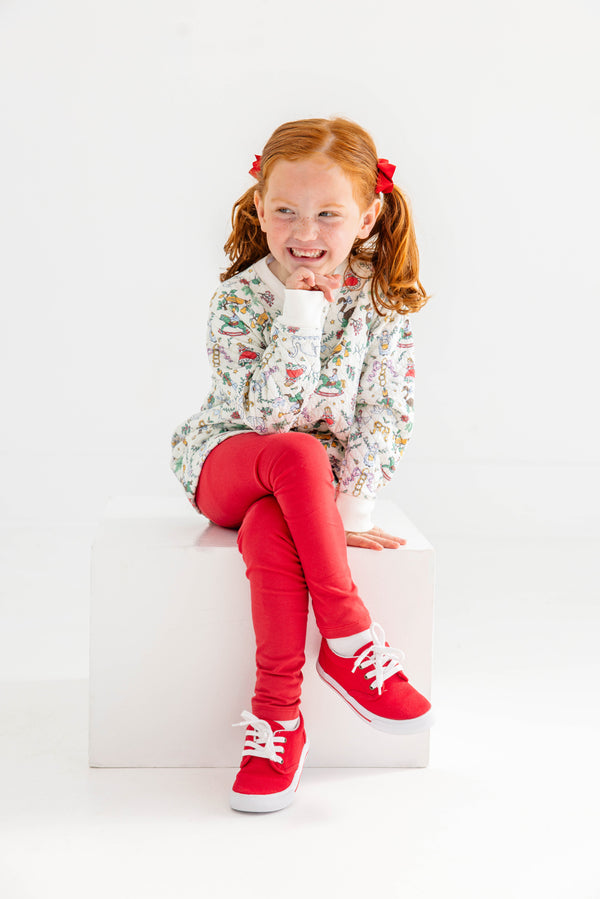 A smiling young girl with red hair in pigtails, dressed in a patterned long-sleeve shirt and The Beaufort Bonnet Company’s Mitzy Sue Slacks in Richmond Red, along with matching red sneakers from the girls’ wardrobe collection, sits on a white cube against a plain white background.