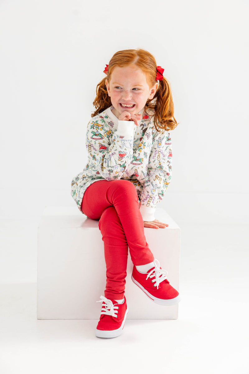 A smiling young girl with red hair in pigtails, dressed in a patterned long-sleeve shirt and The Beaufort Bonnet Company’s Mitzy Sue Slacks in Richmond Red, along with matching red sneakers from the girls’ wardrobe collection, sits on a white cube against a plain white background.