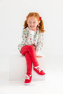 A smiling young girl with red hair in pigtails, dressed in a patterned long-sleeve shirt and The Beaufort Bonnet Company’s Mitzy Sue Slacks in Richmond Red, along with matching red sneakers from the girls’ wardrobe collection, sits on a white cube against a plain white background.