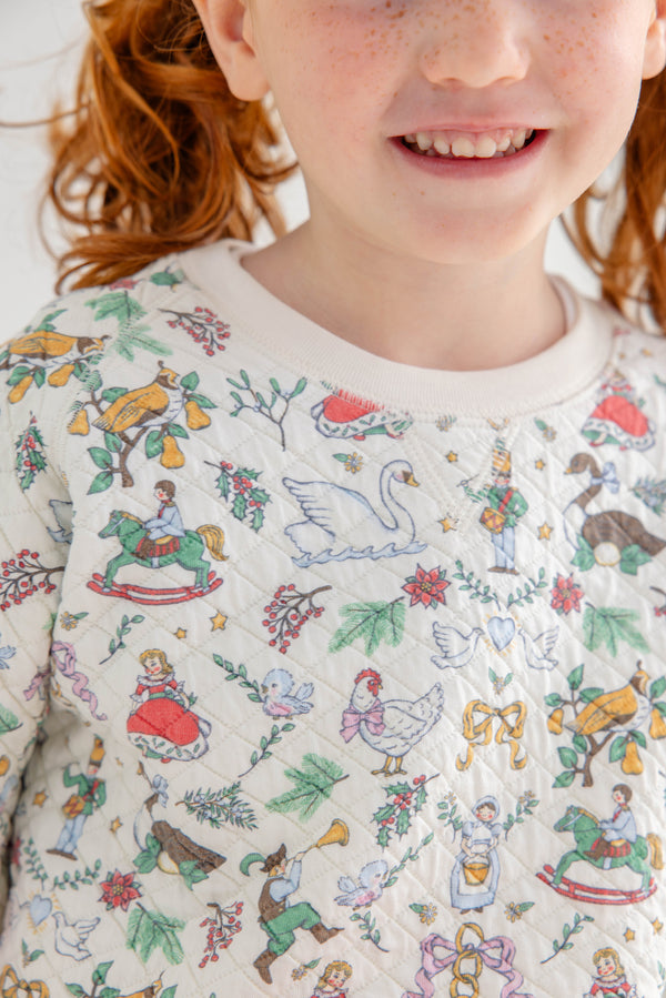 A smiling child with red hair wears The Beaufort Bonnet Company’s Cassidy Comfy Crewneck – 12 Days of Christmas, a quilted sweater adorned with colorful animal, musical instrument, and holiday-themed illustrations. Only the lower half of the child’s face and the top portion of the sweater are visible.