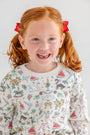 A smiling young girl with red hair in pigtails and red bows wears the Cassidy Comfy Crewneck - 12 Days of Christmas by The Beaufort Bonnet Company, featuring colorful holiday illustrations, set against a plain white background.