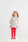 A smiling young girl with red hair in pigtails stands on a white platform, wearing The Beaufort Bonnet Company’s Cassidy Comfy Crewneck - 12 Days of Christmas, paired with red pants, red bows in her hair, and red sneakers, against a plain white background.